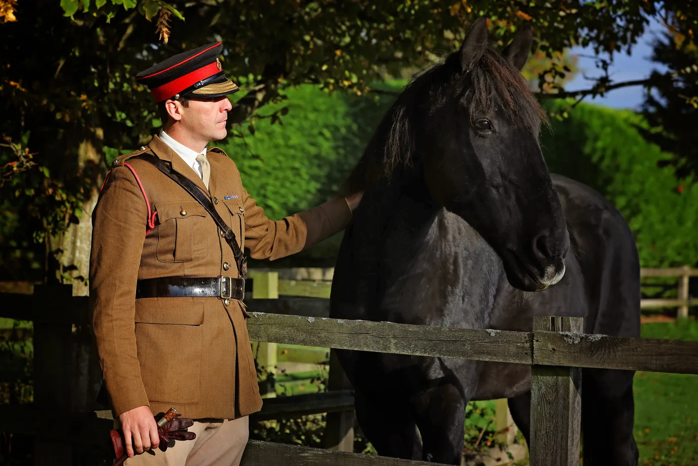 A soldier in Army service dress and a red-banded cap strokes a dark horse over a wooden fence